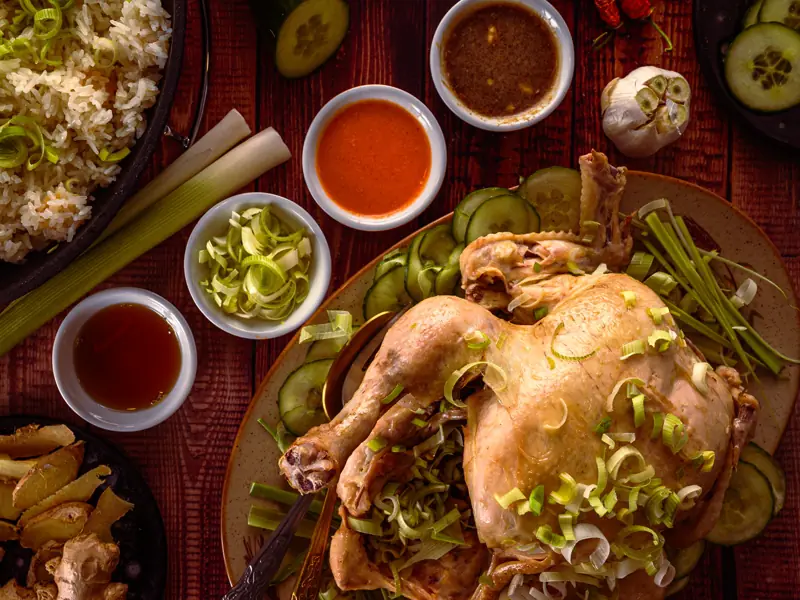 A whole poached chicken on a platter, surrounded by small bowls of chilli, ginger and dipping sauces